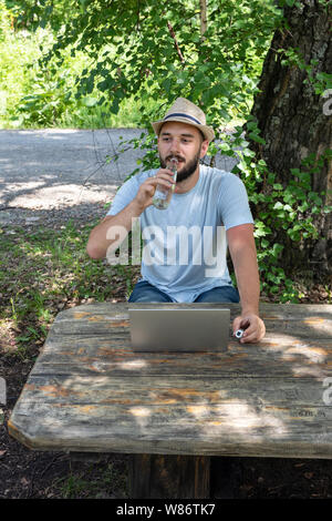 A young caucasian guy, a hipster with a beard, is drinking clear water from a bottle. Sitting in the park with a laptop, tired of long work or study. Stock Photo