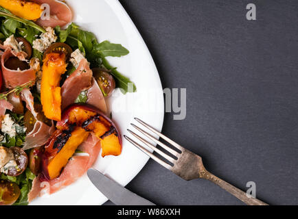 Delicious salad with grilled peaches and prosciutto in a white plate on a black slate background Stock Photo