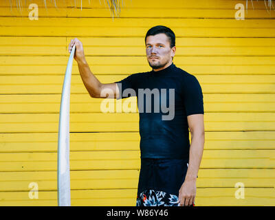 portrait of a surfer on the face of a sunscreen, wearing shorts and a black T-shirt standing with a surfboard. yellow wooden background.