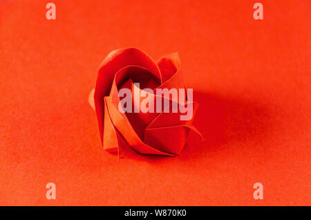 Red origami rose on red background. Japanese art of paper folding. Flat square sheet of paper transferred into a finished sculpture through folding. Stock Photo