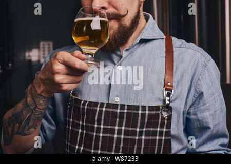 Tattooed man tastes beer from the glass he holds in a close up. Stock Photo