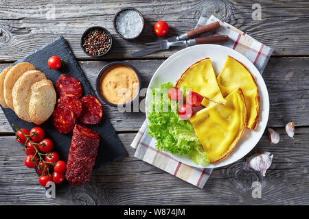 hot omelet on a white plate with lettuce and fresh tomatoes. sliced spicy cured pork sausage and white bread on slate board on a wooden table, view fr Stock Photo
