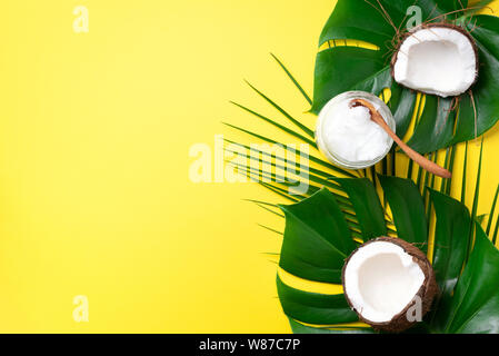 Coconut oil and ripe coconuts, tropical palm and monstera leaves on yellow background with copy space. Top view. Summer creative layout Stock Photo
