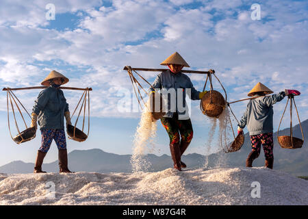 Woman workers pouring freshly harvested salt from baskets to the salt pile at sunrise in Hon Khoi salt field, Nha Trang Province, Vietnam Stock Photo