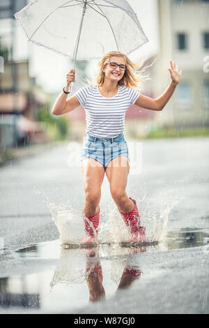 Cheerful girl jumping with white umbrella in dotted red galoshes. Hot summer day after the rain woman jumping and splashing in puddle Stock Photo