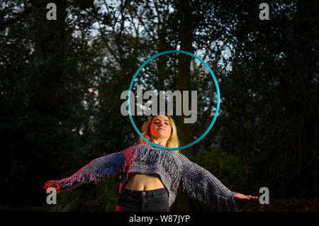 Girl looks directly into the camera through a hoop, which she rolls across her chest, hoop is suspended in mid air and her arms are outstretched, with creative lighting in woods. Stock Photo