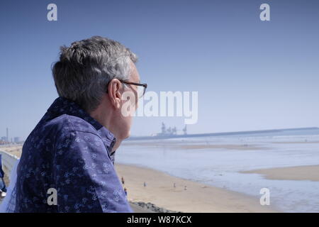 Elderly man looking out over a beach scene, with sand and sea and industrial buildings in the distance Stock Photo
