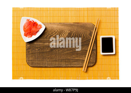 Empty rectangular brown wooden plate with chopsticks for sushi, ginger and soy sauce on yellow bamboo mat background. Top view with copy space for you Stock Photo