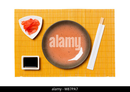 Empty round brown plate with chopsticks for sushi, ginger and soy sauce on yellow bamboo mat background. Top view with copy space for you design. Stock Photo