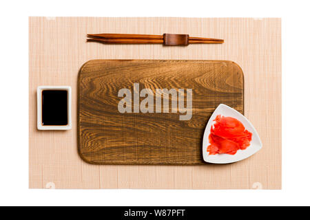 Empty rectangular brown wooden plate with chopsticks for sushi, ginger and soy sauce on brown sushi mat background. Top view with copy space for you d Stock Photo