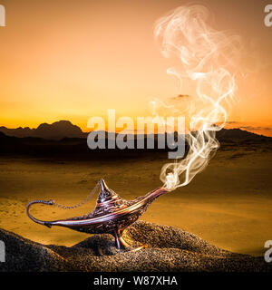 classic gold-colored aladdin lamp laid on the sand of a dune with smoke coming out. desert in the background. Stock Photo