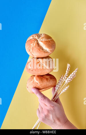 Pyramid of rusty round bread rolls (Kaiser or Vienna rolls), balancing on each other with hand holding wheat ears on blue and yellow colored paper bac Stock Photo