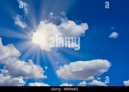 sun is shining bright through white clouds on a blue sky, with sun rays Stock Photo
