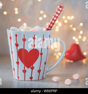 Hot chocolate with marshmallows, red heart on the cup, winter background with lights out of focus. Winter or Valentine's day background. Stock Photo