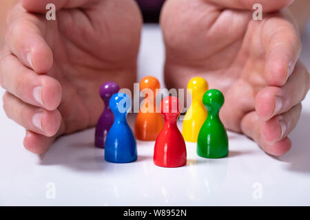 Close-up Of A Person's Hand Protecting Multicolored Pawns Forming Circle Over White Desk Stock Photo