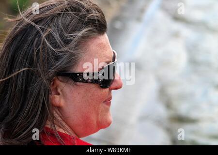Side view of a smiling middle aged white woman with brown hair wearing sunglasses with blurred sea in background. Stock Photo