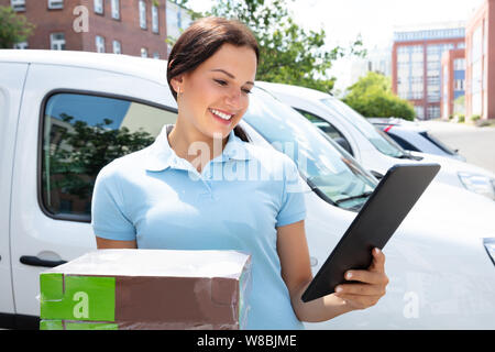 Smiling Delivery Woman Holding Cardboard Box Using Digital Tablet Stock Photo