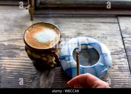 Coffee cigarette on the table. Cigarette in a woman's hand with a beautiful manicure. Blue ashtray. The smoke of cigarettes. Brown cup with coffee on Stock Photo