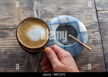 A cigarette with an ashtray and a cup of cappuccino,black coffee with a  cigarette,a tobacco product in an ashtray on a wooden table in a cafe Stock  Photo