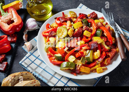 Pisto manchego - Spanish vegetable stew with fried chorizo sausages served on a white plate on a concrete table with sliced bread, view from above, cl Stock Photo