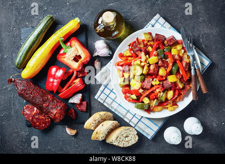 Spanish Pisto manchego - vegetable stew with fried chorizo sausages on a white plate on a concrete table with ingredients, view from above, close-up, Stock Photo