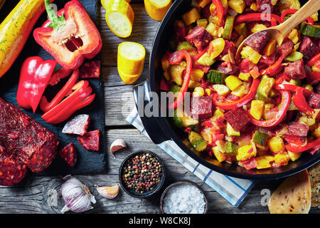 Pisto manchego - vegetable stew with chorizo sausages in a black pan on a rustic wooden table with ingredients on cutting board, spanish cuisine, view Stock Photo