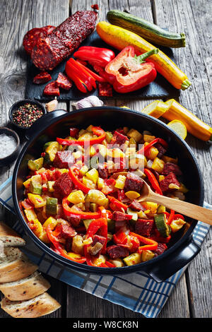 Spanish Pisto manchego - vegetable stew with fried chorizo sausages in a black pan on a rustic wooden table with ingredients, vertical view from above Stock Photo