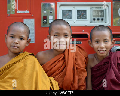 Three Burmese Buddhist boy monks with robes of three different colours pose for the camera in front of a red fire truck. Stock Photo