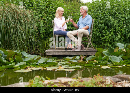 Mature  Couple Celebrating With Champagne Sitting On Chairs On Wooden Jetty By Lake Stock Photo