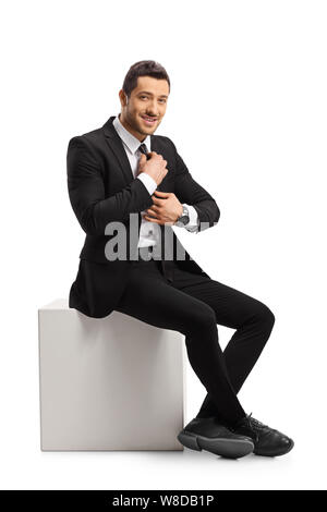 Full length portrait of a young man in a suit sitting and fixing his tie isolated on white background Stock Photo