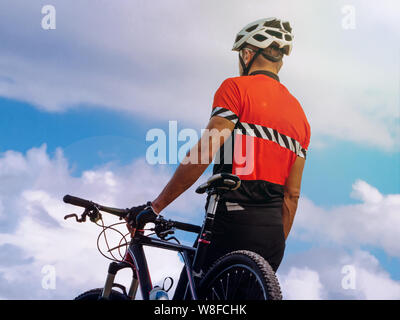 Concept cyclist stands on the top of a mountain near a mountain bike, background is a blue sky with clouds. back view. Toned. Stock Photo