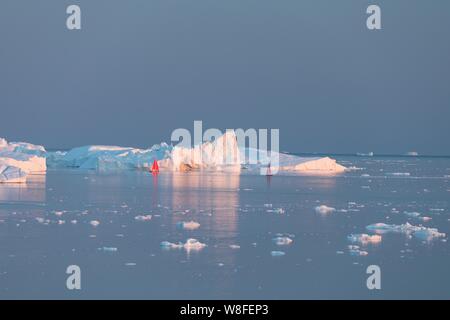 Little red sailboat cruising among floating icebergs in Disko Bay glacier during midnight sun season of polar summer. Ilulissat, Greenland  Expedition Stock Photo