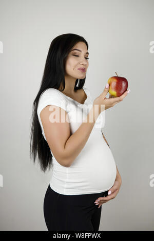 beautiful pregnant brunette woman holding red fresh apple smiling standing half a turn on isolated white background Stock Photo