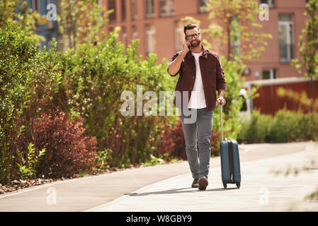 Happy to hear you Cheerful bearded man in casual wear and eyeglasses pulling his luggage and talking by phone while walking through the city street. Travel concept. Lifestyle Stock Photo