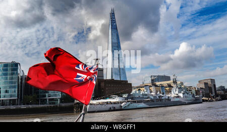 The London Shard and HMS Belfast with Clipper River Boat Red Ensign Marine Flag flying in the breeze, with dramatic sky viewed from Thames Clipper RB1 river boat with ‘red duster’ flag flying. HMS Belfast, The Shard and More London Place South Bank London SE1 UK