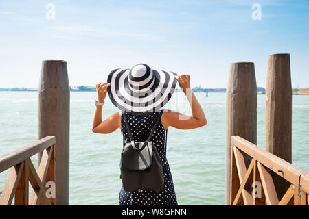 Rear View Of Woman With Her Backpack Standing On Wooden Pier Looking Over The Bay Stock Photo