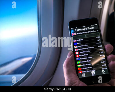 Currency Exchange Rates screen display at historic rates 2015  UK Pound to Dollar, Euro, Swiss Franc, Chinese Yuan etc., Hand holding iPhone smartphone in aircraft cabin with window, wing and sky beyond. Stock Photo