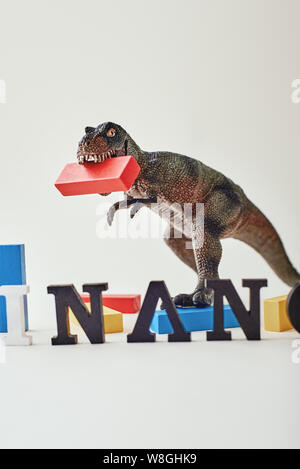 Close up of red, blue and yellow wooden blocks placed unevenly over light background near dinosaur toy. Word Finance made of wooden letters standing in a row. Vertical shot Stock Photo