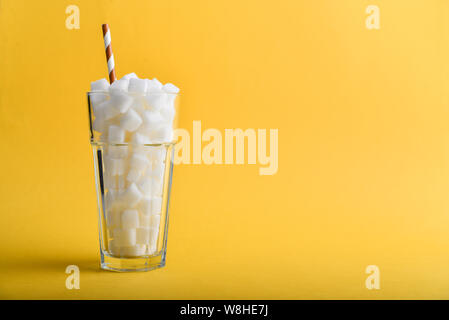 Glass full of sugar cubes with drink straw on yellow background - unhealthy diet concept. Stock Photo
