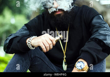 Man with a beard smokes an electronic cigarette sit on a bench in the park Stock Photo