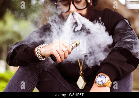 Man with a beard smokes an electronic cigarette sit on a bench in the park Stock Photo