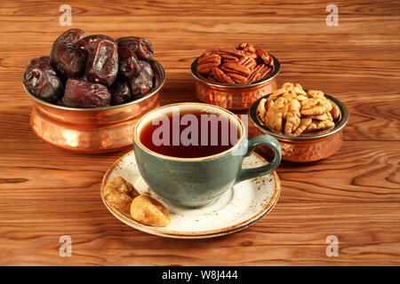 Vegetarian dessert: dried dates, pecans and walnuts in copper bowls, dried figs on a saucer and a cup of black tea on a brown wooden table. Closeup Stock Photo