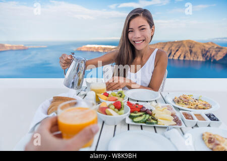 Couple eating breakfast. Smiling tourist woman drinking coffee and man drinking orange juice on terrace resort outdoor. Healthy and delicious food served for breakfast. Santorini, Greece. Stock Photo