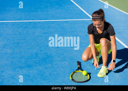 Sports woman Asian happy athlete getting ready for playing tennis tying laces of her running shoes on outdoor blue hard court. Professional player preparing for summer tournament game. Stock Photo