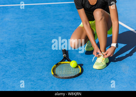 Sports woman athlete getting ready for playing a game of tennis tying laces of her running shoes lacing the shoelaces on outdoor blue hardcourt in summer. Professional player preparing for tournament. Stock Photo