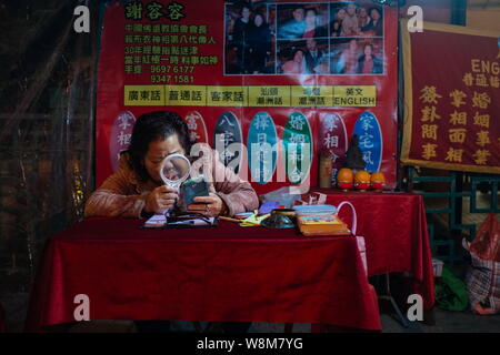 A fortuneteller uses her smartphone with a magnifying glass at her stall in Yau Ma Tei, Hong Kong, China, January 2016.   With the impact of internati Stock Photo