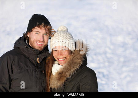 Happy winter interracial couple outdoors portrait. Cute young adults smiling in warm outerwear knit hat and down coats outside. Asian chinese woman, Caucasian man. White snow ice background texture. Stock Photo