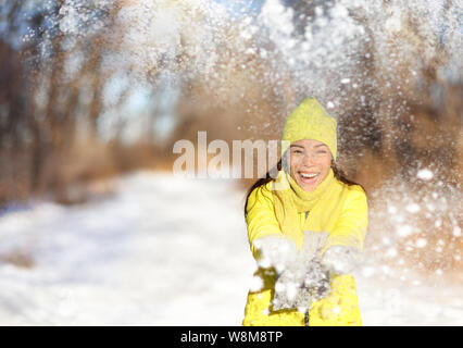 Winter snow fight happy girl throwing snow playing outside. Joyous young Asian woman having fun in nature forest park on snowy day wearing yellow outerwear with warm accessories: gloves, hat, scarf. Stock Photo