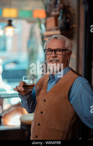 Grey-haired man wearing brown vest drinking whisky Stock Photo