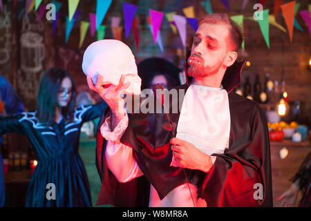 Attractive young man dressed up like dracula holding a black rose looking at his human skull at halloween celebration. Dracula costume. Stock Photo